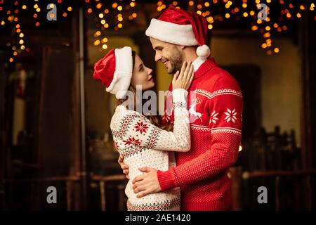 Happy young couple in santa hats et chandails chauds embrassant tout en regardant les uns les autres Banque D'Images