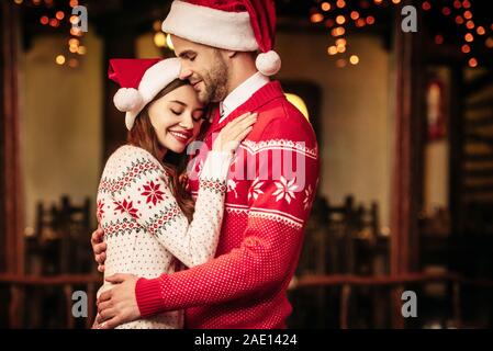 Happy young couple in santa hats et chandails chauds embrassant les yeux fermés Banque D'Images