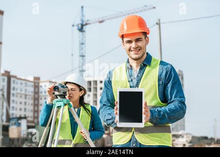 Arpenteur Smiling holding digital tablet avec écran vide et collègue à la mesure de niveau à l'arrière-plan Banque D'Images