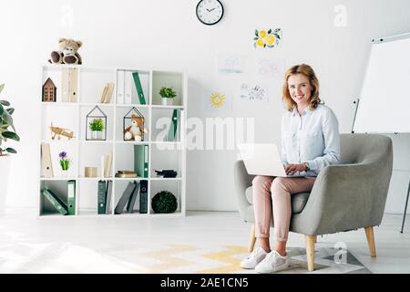 Jolie et souriante en psychologue pour enfants shirt holding laptop et assis sur un fauteuil Banque D'Images