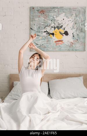 Happy girl sitting in bed with closed eyes et d'étirements avec main levée Banque D'Images