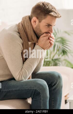 Homme malade en écharpe toux en mains jointes tandis que sitting on sofa Banque D'Images