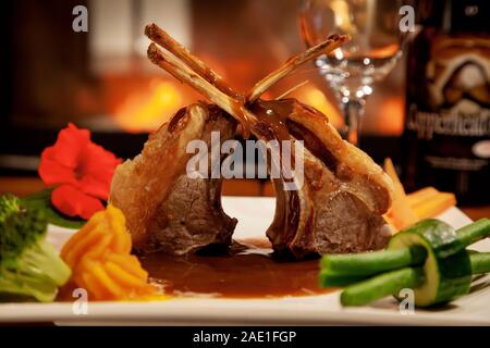 Close up sur des côtelettes d'agneau rôti avec des légumes et de la sauce sur une plaque blanche avec du vin d'accompagnement et d'une cheminée Contexte Dans le cadre d'un restaurant. Banque D'Images