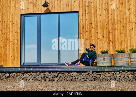 Jeune homme en tablier se trouve sur la terrasse d'une maison en bois. Planche de bois jaune. Fenêtres panoramiques. Parfait reflet dans une fenêtre de ciel bleu. La conc Banque D'Images