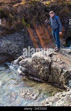 Homme debout sur des rochers holding Panasonic caméra vidéo pour capturer le paysage de conte de piscines, river cassante, Isle of Skye, Scotland, UK en Mars Banque D'Images