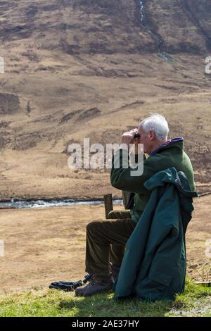 Homme assis sur un banc regardant à travers des jumelles à Fairy Piscines, Isle of Skye, Scotland, UK en Mars Banque D'Images