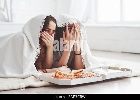 Drôle et peur. Couverture chaude. Soeurs de manger une pizza en regardant la télévision tout en se trouvant sur l'étage de belle chambre dans la journée Banque D'Images