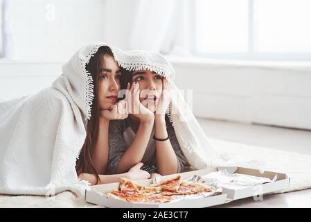 C'est incroyable. Couverture chaude. Soeurs de manger une pizza en regardant la télévision tout en se trouvant sur l'étage de belle chambre dans la journée Banque D'Images