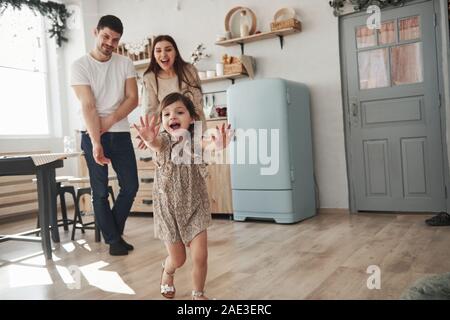 Elle va vous donner un câlin. Enfant de sexe féminin ludique s'amuser en exécutant dans la cuisine dans la journée d'avant de sa mère et de son père Banque D'Images