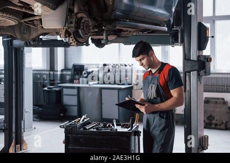Être précis avec les chiffres. L'homme à l'atelier en uniforme à l'aide du bloc-notes pour son travail de fixation voiture cassée Banque D'Images