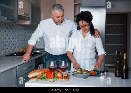 Faire une salade. L'homme et sa femme en chemise blanche à la préparation de la nourriture sur la cuisine en utilisant des légumes Banque D'Images