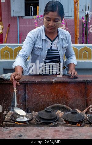 Une jeune femme birmane verse la pâte pour faire une crêpe traditionnelle (ah l'arrière-boutique) sur un feu ouvert unique cuisinière avec couvercles dans une ville rurale au Myanmar (Birmanie) Banque D'Images