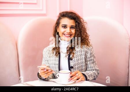 Jeune femme assise dans un café à la table en bois, à boire du café et à l'aide de téléphone. Banque D'Images