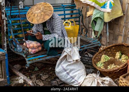 Un homme dans un chapeau de riz conique ou coolie hat se détend sur un banc bleu avec un téléphone cellulaire après l'achat de pommes dans le marché ferroviaire de Mandalay, Myanmar Banque D'Images