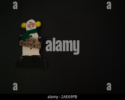 En bois peint ornement bonhomme vert avec foulard et cache-oreilles jaune tenant une 'Snow arrive' signe sur un fond noir. Banque D'Images