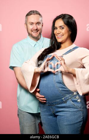 Heureux homme et femme enceinte en salopettes en denim sur fond rose vide en studio Banque D'Images