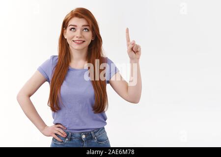 Jolie rousse de rêve fille moderne avec de longs cheveux rouges, de rechercher des curieux, vers le haut, intrigué, souriant comme départ amusant, signe de lecture promo Banque D'Images