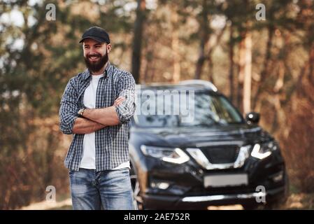 Sourire pour la caméra. Homme barbu près de sa toute nouvelle voiture noire dans la forêt. Concept de vacances Banque D'Images