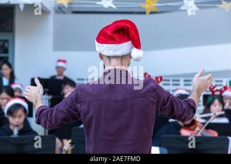 Vue arrière d'un homme d'orchestre, le port de tenue décontractée avec rouge santa hat, mener son jeune groupe jouant de la musique de Noël à un événement en plein air, hol Banque D'Images