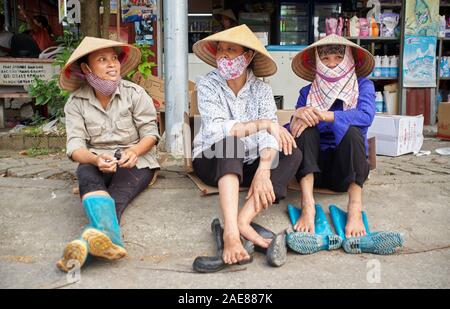 Vietnam, bac Ninh, trois femmes portant des chapeaux coniques traditionnels et des masques faciaux s’assoient sur des boîtes en carton au bord de la route, prenant une pause de leur travail Banque D'Images