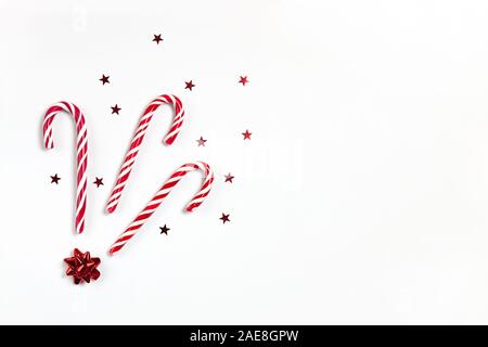 Composition de Noël trois cannes de bonbon au caramel, confettis étoiles et arc rouge sur fond blanc avec l'exemplaire de l'espace. Style minimal de mise à plat de fête. Pour g Banque D'Images