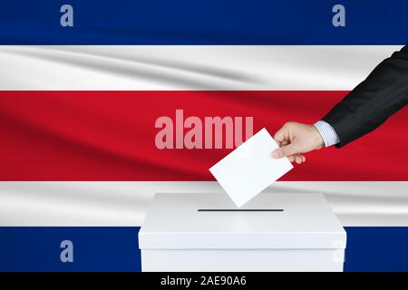 Élection au Costa Rica. La main de l'homme qui a mis son vote dans l'urne. Drapeau du Costa Rica ondulé sur fond. Banque D'Images