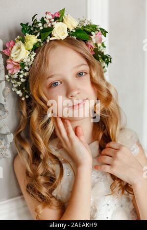 Portrait d'une jolie petite fille aux cheveux blonds dans une robe rose est assis dans un studio lumineux avec une couronne de fleurs sur sa tête. Banque D'Images