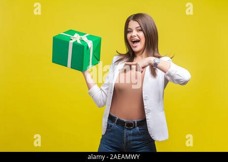 Portrait de femme brune en veste et jeans pointant sur boîte-cadeau dans sa main, avec la bouche ouverte et de surprise, agréablement s Banque D'Images