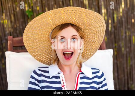 Closeup portrait of happy laughing woman inopinément en robe et chapeau assis sur un transat confortable et à la recherche à l'appareil photo avec une bouche ouverte Banque D'Images