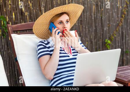 Portrait de la belle jeune femme d'adultes en étonné hat et robe assis sur chaise longue avec ordinateur portable et faire appel avec smartphone, avec surprise f Banque D'Images