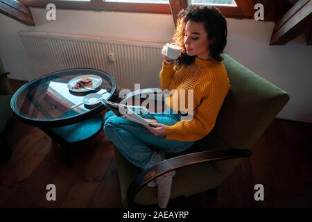 Jeune femme séduisante en jaune chandail confortable assis sur un fauteuil vert, boire du thé chaud et de la lecture à la Mansart Grenier Magazine. Concept de vie lente Banque D'Images