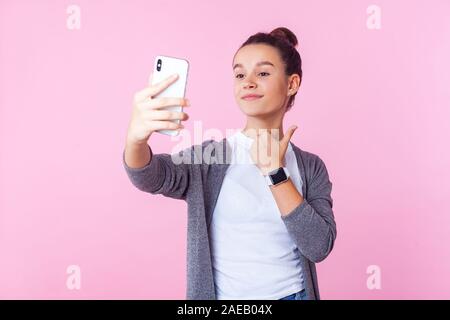 Comme ! Portrait of teenage girl with positive bun hairstyle dans les tenues de sourire et showing Thumbs up tout en faisant appel vidéo, prendre sur selfies p Banque D'Images