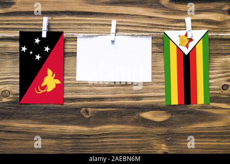 Drapeaux suspendus de la Papouasie-Nouvelle-Guinée et le Zimbabwe attaché à corde avec pinces à linge avec de l'espace de copie sur papier blanc sur fond de bois diplomatique. Banque D'Images