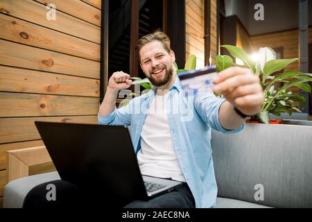 Attractive smiling Caucasian man using laptop in co-working space, holding credit card et effectuer un paiement en ligne. Paiement en ligne avec succès. Banque D'Images