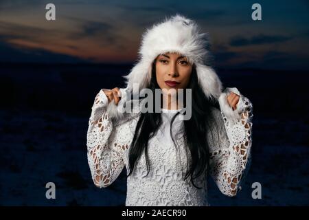 Belle jeune fille avec de longs cheveux noirs sur fond de ciel du soir et le champ de neige. Modèle de charme vêtu de blanc blouse en tricot blanc toucher chapeau chaud. Banque D'Images