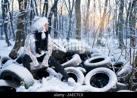 Belle fille assise sur de vieux pneus usagés couverte de neige déversées en milieu de forêt. Model wearing hat en bleu et blanc, chemisier blanc, jeans et baskets à l'hôtel. Banque D'Images