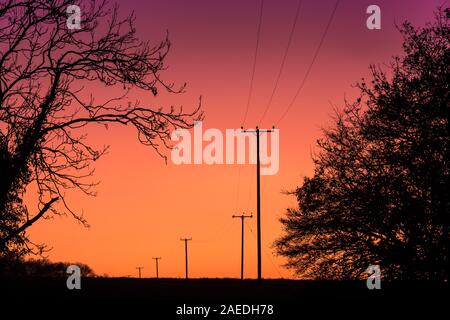 Les pylônes d'électricité du réseau national rural transportant des lignes électriques traversent la campagne du Suffolk, East Anglia, Royaume-Uni après un coucher de soleil hivernal. Banque D'Images