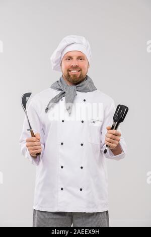Jeunes chef barbu en tenue uniforme en vous regardant et ustensiles Banque D'Images
