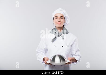 Happy young female chef réussie en uniforme portant cloche avec repas maison Banque D'Images