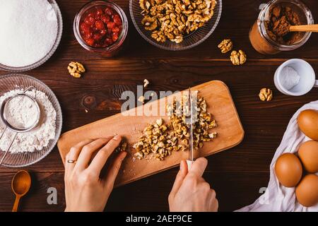 Cuisine maison, processus de mise à plat. Woman's hands hacher les noix pour la cuisson du gâteau brownie parmi les ingrédients sur la table en bois sombre, vue du dessus. Banque D'Images