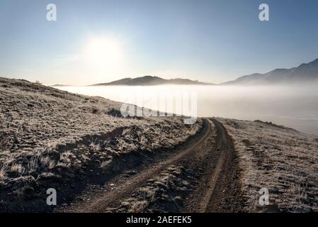 Paysage d'automne étonnant avec un chemin de terre dans les montagnes, un épais brouillard blanc moelleux sur le lac, de l'herbe dans le givre et le soleil contre un ciel bleu à Banque D'Images