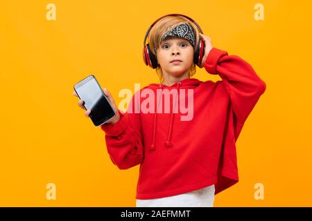 Garçon blond européen dans une capuche rouge est à l'écoute de la musique au casque rouge et est titulaire d'smartphone avec immersive sur un fond orange Banque D'Images