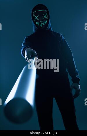 Portrait d'un criminel anonyme homme avec batte de baseball dans un hoodie noir cachant son visage derrière un masque de néon. Angle inférieur Banque D'Images