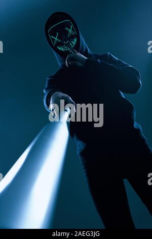 Portrait d'un criminel anonyme homme avec batte de baseball dans un hoodie noir cachant son visage derrière un masque de néon. Angle inférieur Banque D'Images