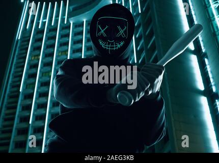 Portrait d'un criminel anonyme homme avec batte de baseball dans un hoodie noir cachant son visage derrière un masque de néon. Arrière-plan de ville lumineuse Banque D'Images