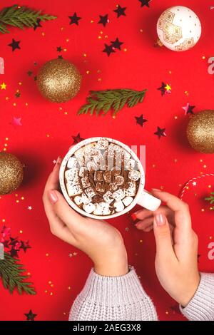 Woman's hand holding white mug cacao de Noël avec des guimauves sur un fond rouge parmi des décorations de Noël. Haut de la vue, télévision lay. Banque D'Images