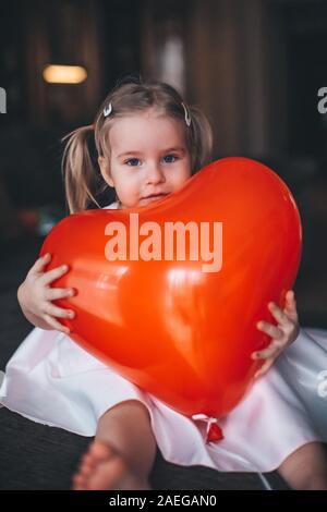 Adorable petite fille blonde assise sur un cheval de bois foncé et de table en forme de coeur rouge étreindre ballon à la maison. Concept de la Saint-Valentin Banque D'Images