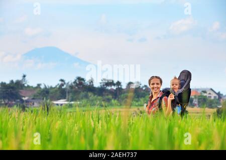 Promenade dans le champ de riz vert. Heureux mère peu voyageur en exerçant son sac à dos. Baby ride sur femme dos. Voyage d'aventure, la randonnée avec des enfants Banque D'Images