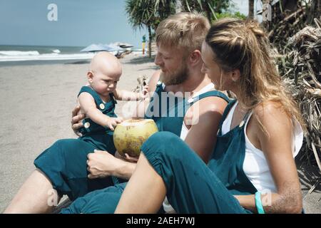 Drôle de famille heureux hipster pique-nique sur plage - mère, père nourrir bébé garçon. Petit-fils de manger des fruits avec plaisir, boire de noix de coco fraîche. Mode de vie sain, l'act Banque D'Images