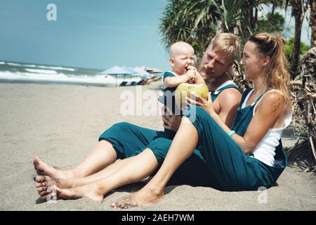 Drôle de famille heureux hipster pique-nique sur plage - mère, père nourrir bébé garçon. Petit-fils de manger des fruits avec plaisir, boire de noix de coco fraîche. Mode de vie sain Banque D'Images